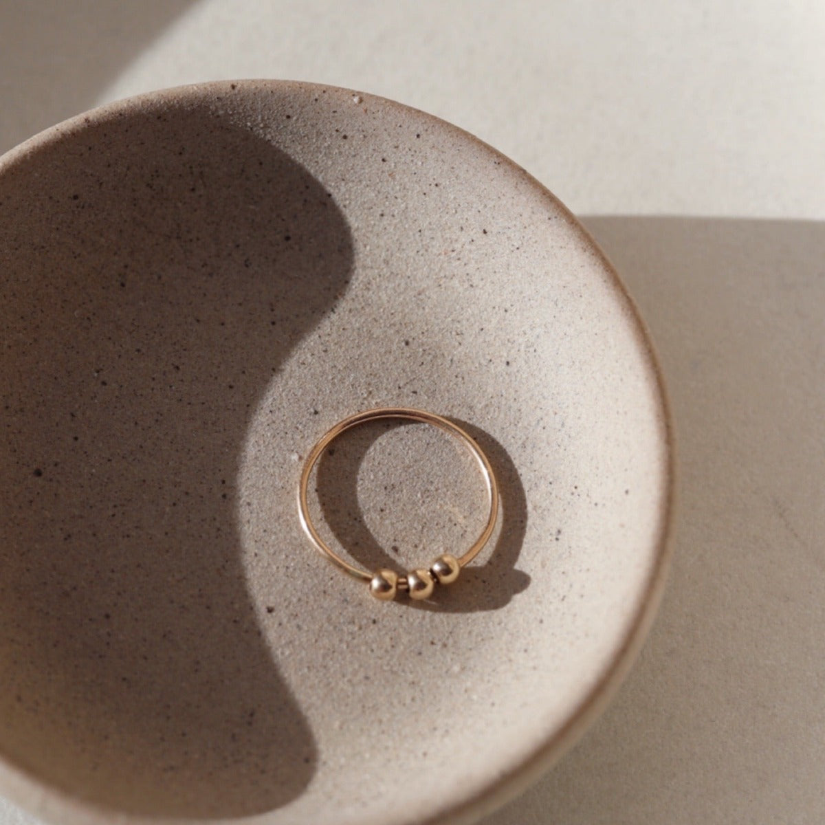 14k gold fill Meridian Fidget Ring placed on a gray plate in the sunlight. This ring features alleviate anxiety or improve focus. Each ring is adorned with 3 gold filled beads that spin and roll so you can play with the ring as needed. - Token Jewelry