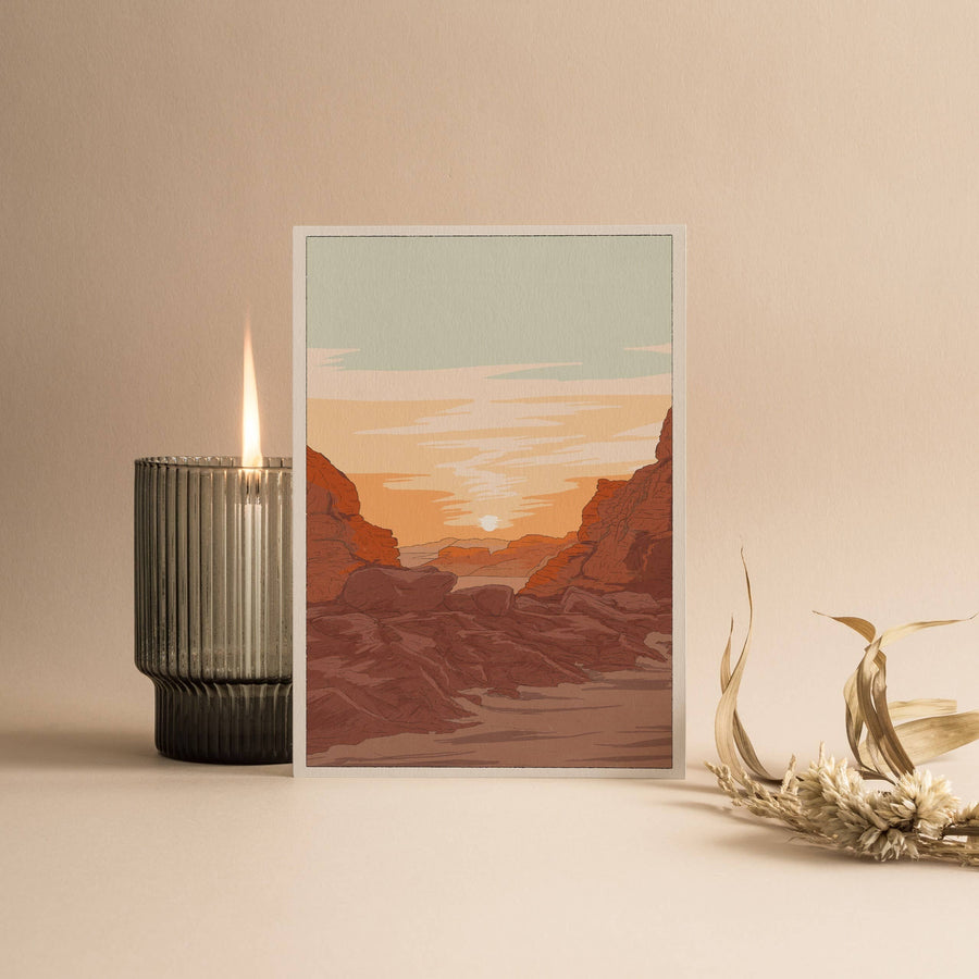 Candle with a card with a view of an overlook of the Utah desert with a sunset in the background. As well as a flower sitting along the card.