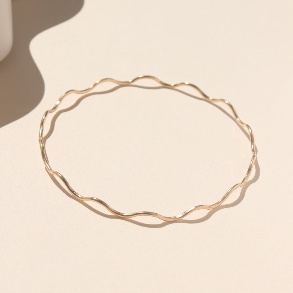 14k gold fill bangle with wave-like textured wire, displayed on a cream backdrop in natural sunlight.