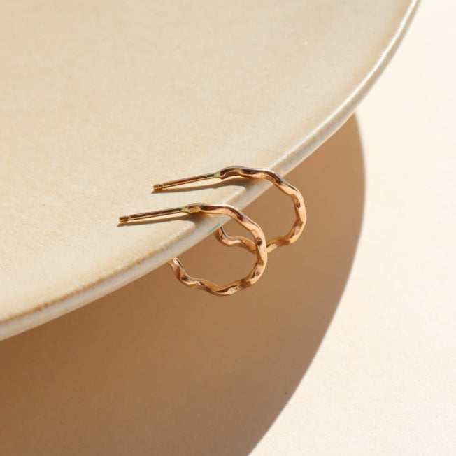 A wave like 14k gold fill textured huggie earring, displayed on a tan dish in the sunlight.