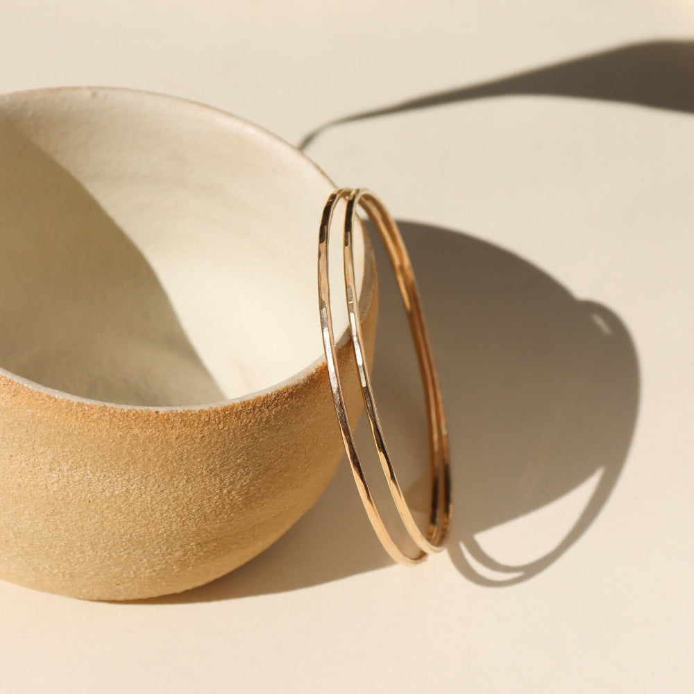 Fine Line Bangle in gold fill and sterling silver, hammered to a shine, shown stacked with other bracelets and worn solo for a minimalist look.
