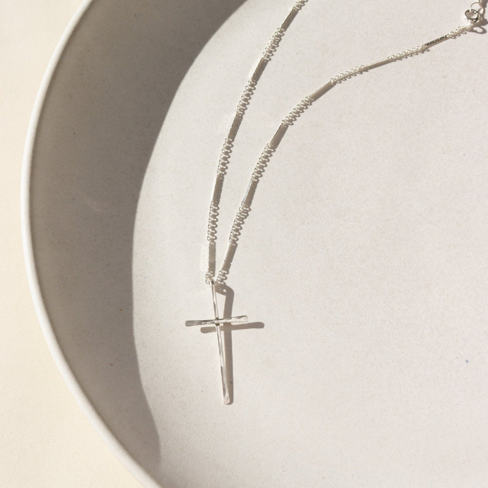 A sterling silver thin bar and link chain featuring a 1 1/2" hammered cross pendant. The necklace is displayed on a white dish.
