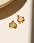 Gold pendant charms featuring aventurine and rose quartz cabochons in organic gold settings, displayed on a neutral surface in warm natural light.