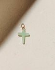 Chrysoprase cross charm featuring a soft green gemstone cross wrapped with gold wire, photographed on a warm neutral surface with soft natural light and shadows.