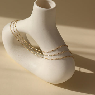 14k gold fill Hera Choker placed on a white pot set in the sunlight. This necklace features three layers of chains making it look like you're wearing three necklaces.