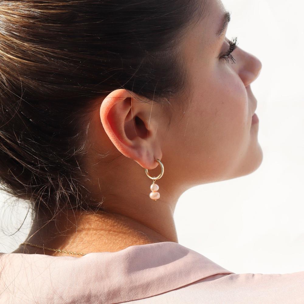 Pink pearl earrings are taken on a brunette model wearing a light pink blouse.