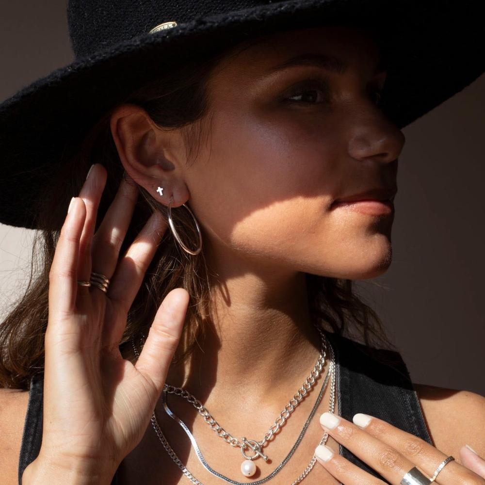 Model wearing Sterling silver cross stud earrings. Along with other sterling silver jewelry. Model is wearing a black denim top with a black hat.