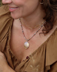 Oval cream, lavender, and golden stone pendant on a multicolored beaded necklace, layered with a delicate gold chain and worn with a brown top.