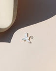 A small iridescent mother-of-pearl bird charm with a sterling silver loop, photographed on a cream-colored surface in sunlight, casting a soft shadow.