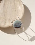 The siren cuff features a Terra Agate Gemstone wire wrapped to a sterling silver wide cuff. The cuff is displayed against a cream plate in the sunlight.
