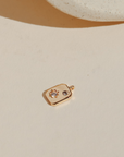 Mini gold celestial charm featuring a sparkling sun and moon design on a rectangular tag, photographed on a neutral surface in warm natural light.