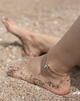 14k gold fill anklet with a swivel push-gate clasp holding a seashell charm and a turquoise charm, photographed on a model with feet in the sand