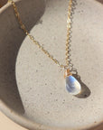 14k gold fill necklace with natural teardrop moonstone full of blue flash laid on a gray moonstone plate.