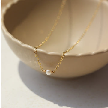 14k gold fill Mini Pearl Necklace laid on a tan bowl in the sunlight. This Necklace features a mini pearl with connected by the simple chain.