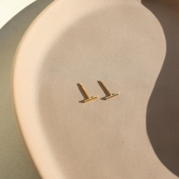 14k gold fill bar studs that are laid across a peach plate that is laying in the sun.