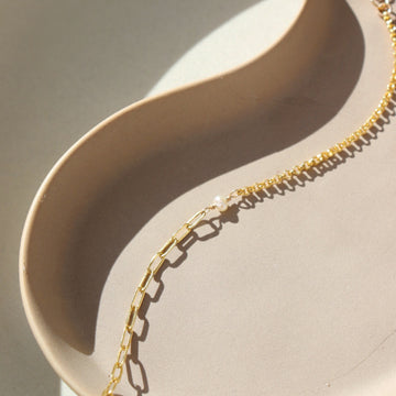 14k gold fill bracelet place on top of a peach colored plate. Sitting in the sunlight, featuring the narrow link chain, a tiny pearl gemstone, and the Dylan chain. The jewelry is handmade in Eau Claire Wisconsin, at Token Jewelry.