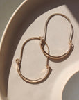 14k gold fill hammered Paloma hoops, earrings are placed on a peach colored plate in the sunlight. These earrings feature a wire around 1/2 inch shaped like an oval, then followed by the other half of the oval that is hammered. 