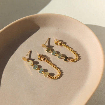 14k gold fill Starlight Chain Studs placed on a tan plate in the sunlight. This earring features the polka dot stud with a dangle of the starlight chain along with the la mer chain. Making this the perfect every day earring.