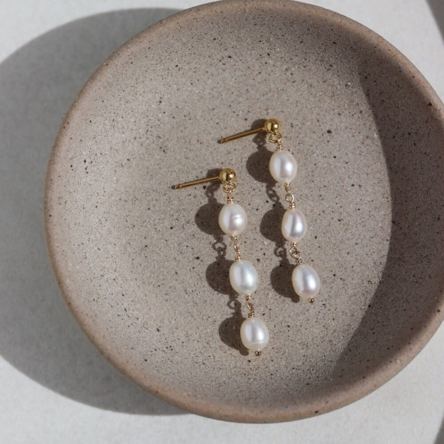 14k gold fill Trillium Pearl Drop Earrings laid on a gray plate. These earrings a perfect stud earring that gives the dangle look.