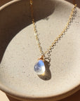 14k gold fill necklace with natural teardrop moonstone full of blue flash laid on a gray moonstone plate.