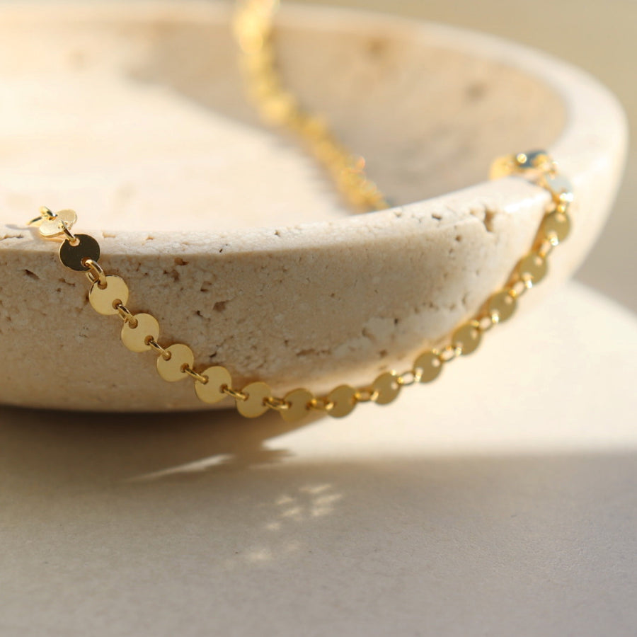 14k gold fill Starlight Choker laid in the sunlight on a gray plate. This choker is perfect for layering. This Necklace features circle disc that are linked together.