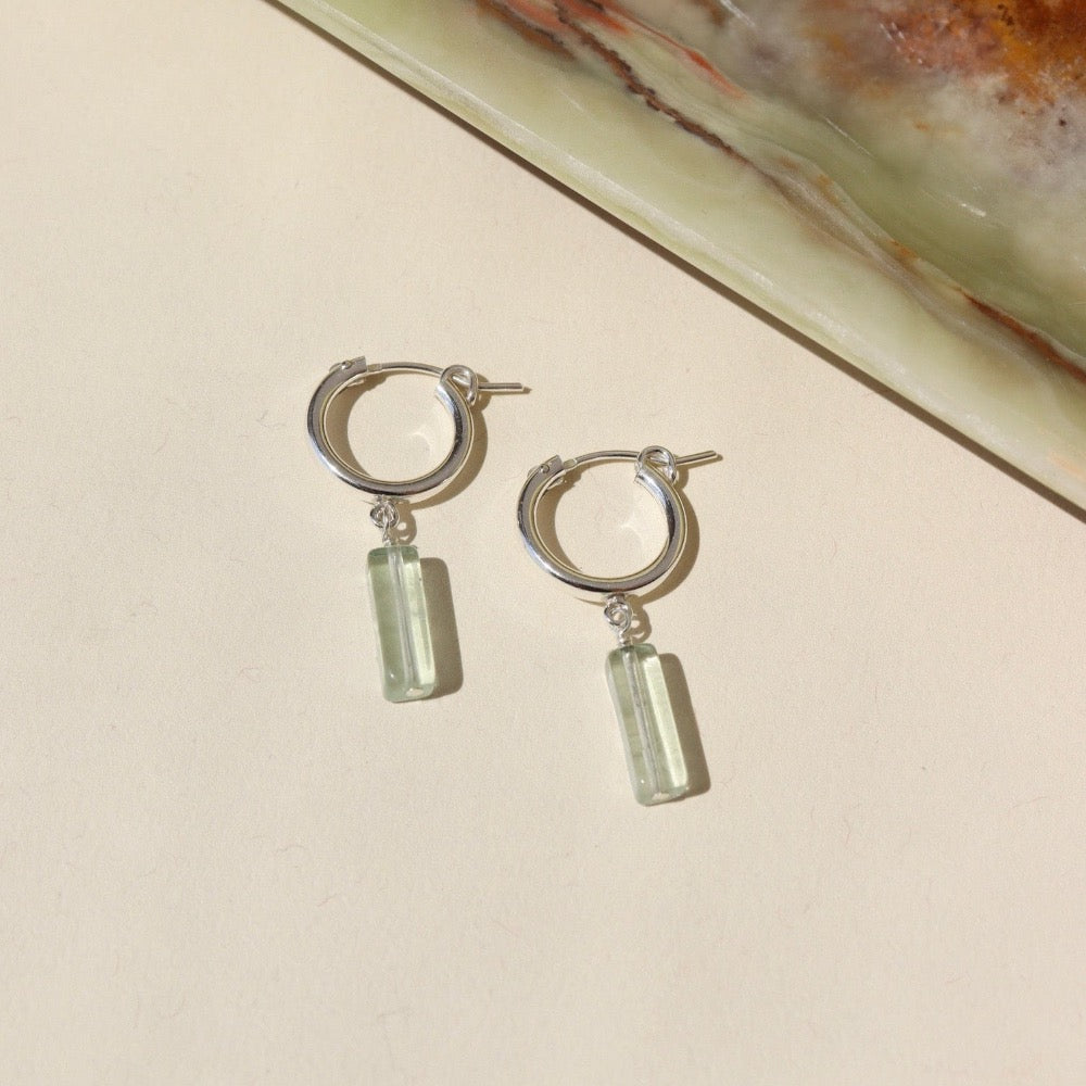 14k gold fill Flynn Hoops laid on a tan plate in the sunlight. These earrings feature a hoop like earring with a dangle of a Fluorite tube gemstone.