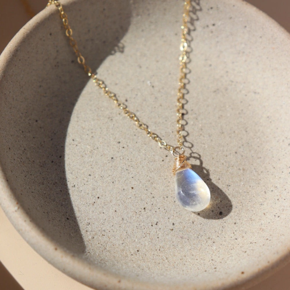 14k gold fill necklace with natural teardrop moonstone full of blue flash laid on a gray moonstone plate.