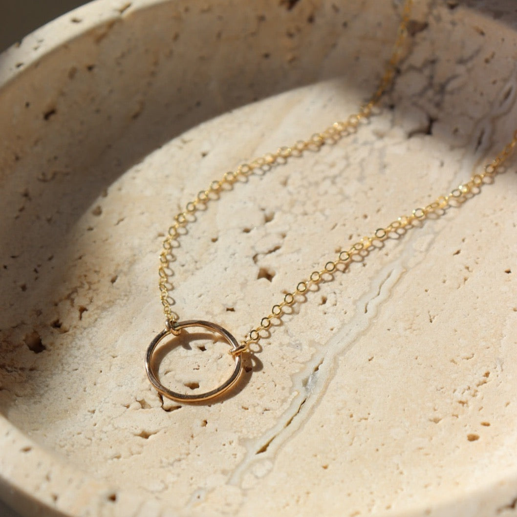 14k gold fill Eternity Necklace placed on a cream colored dish. This necklace and plate is sitting in the sunlight. This necklace features an open circle disc that is attached to our simple chain. Token Jewelry is hypoallergenic and nickel free. - Token Jewelry