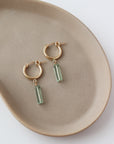 14k gold fill Flynn Hoops laid on a tan plate in the sunlight. These earrings feature a hoop like earring with a dangle of a Fluorite tube gemstone.