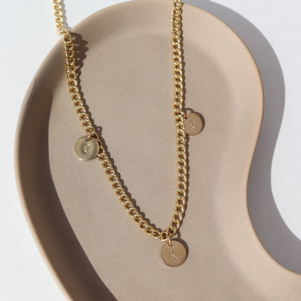 14k gold fill Modern Muse Initial Necklace laid on a tan plate in the sunlight. This Necklace feature our Demi Alexandra chain featuring a charm initial of your choice.