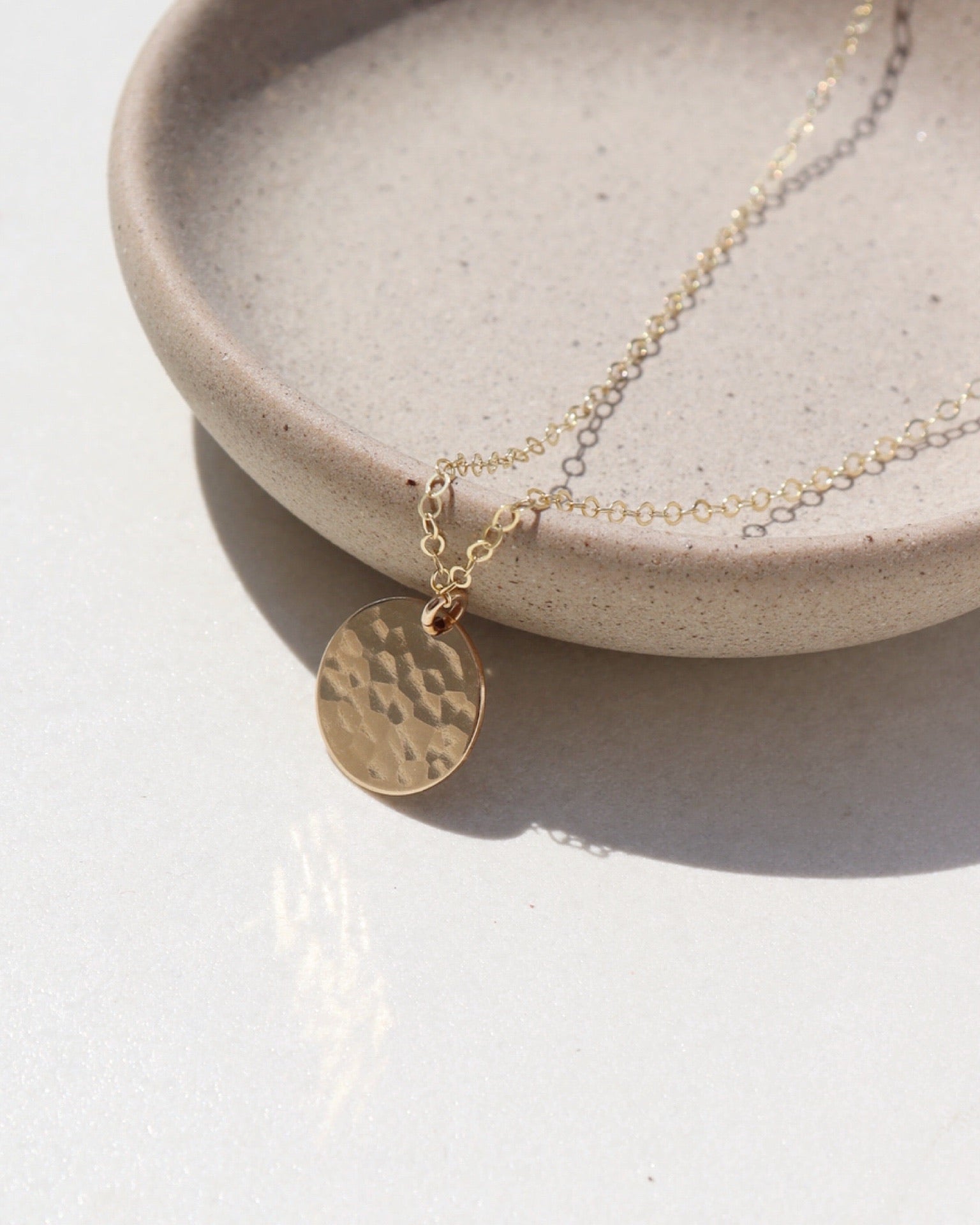 14k gold fill Moon drop necklace laid on a plate in the sunlight. This necklace features our simple chain met by a hammered coin, making it look like the moon.