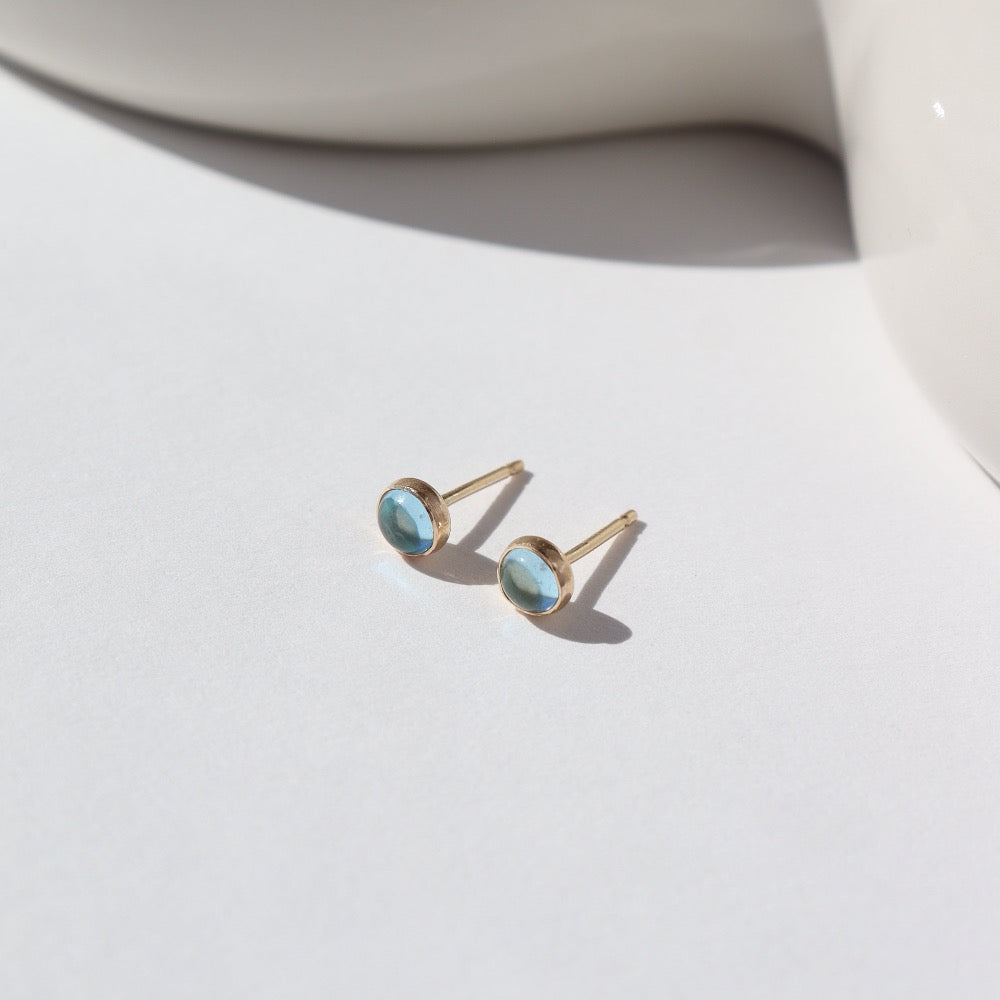 4mm Blue Topaz gemstones set in a 14k gold fill bezel stud earring and placed on a white plate for display. Sold as a pair and perfect for single or double pierced ears.