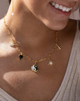 14k gold fill chain with various gold charms, featuring a Butterfly, black heart, yin yang symbol, opal star, and a fleur de lis, photographed on a model wearing a beige knit tank top