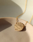 14k gold fill full moon necklace laid on a peach colored plate.  Necklace is laid in the sunlight. This necklace features the full moon disc that is hammered in the middle and connected by the simple chain. 