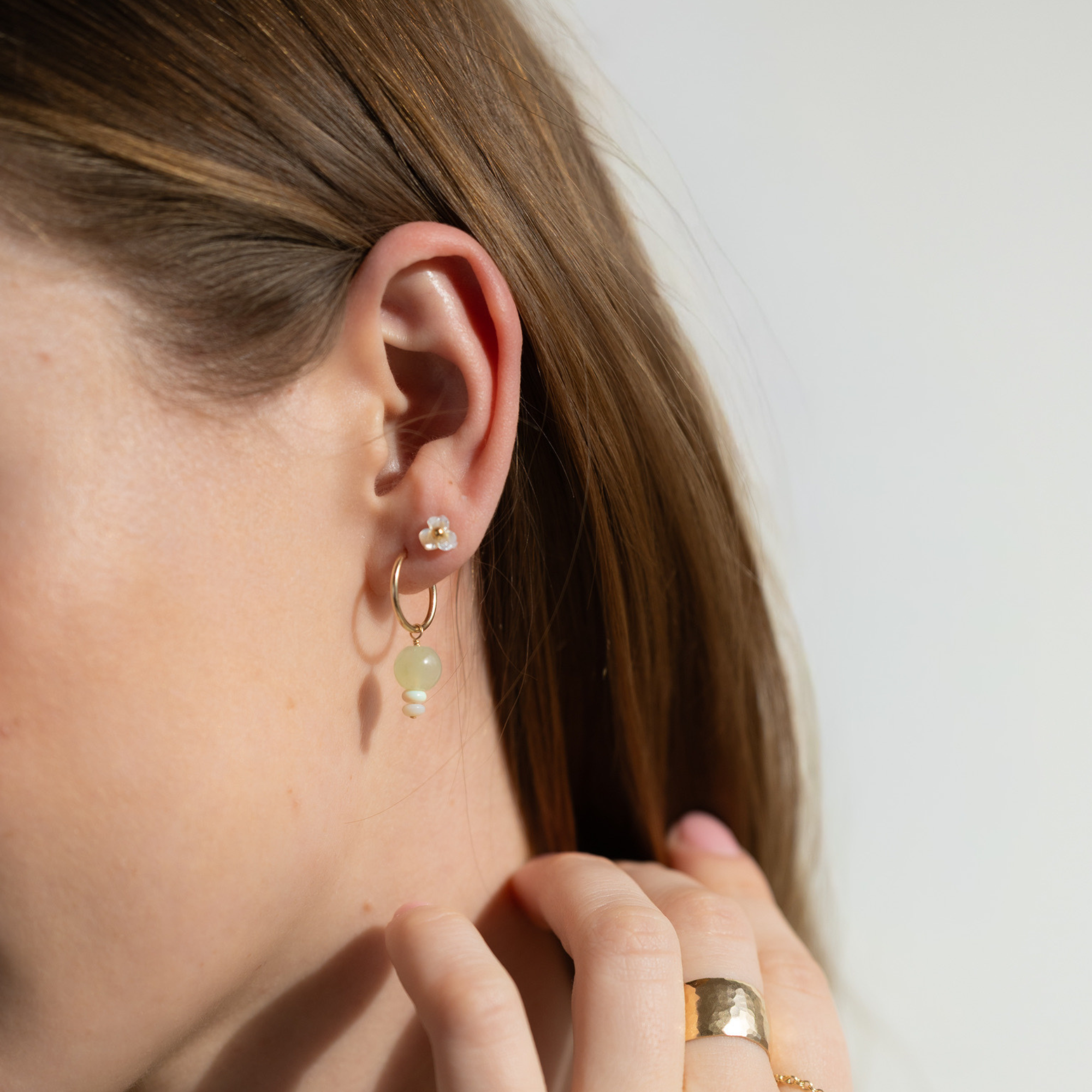 Model wearing small mother-of-pearl flower stud earrings with a polished gold center, styled alongside gold hoop earrings and a green gemstone drop earring.
