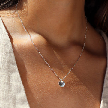 a sterling silver necklace featuring a dainty chain and a small, concave, polished disc, photographed on a model wearing a cream colored linen vest