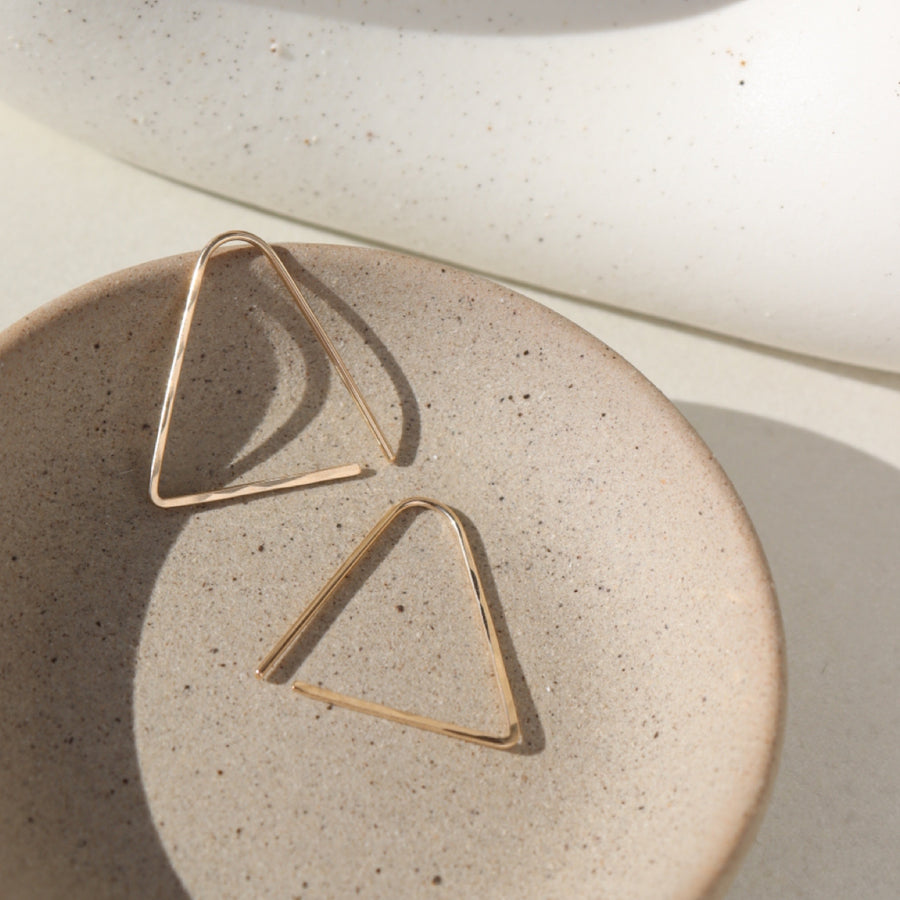 14k gold fill Sunday Earrings laid on a gray plate in the sunlight. These earrings feature a triangle look easy to thread in your ear.