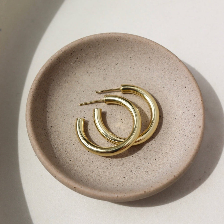 14k gold fill Harper Hoops laid on a tan paper in the sunlight. These earrings feature a classic smooth hoop with a post making the easy to take in and out.