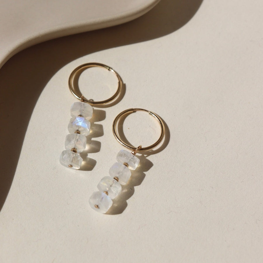 14k gold fill Selene Hoops laid on a tan paper in the sunlight. These Earrings feature our 16mm Goldie hoops along with 4 Moonstone Faceted gemstones stacked onto of each other