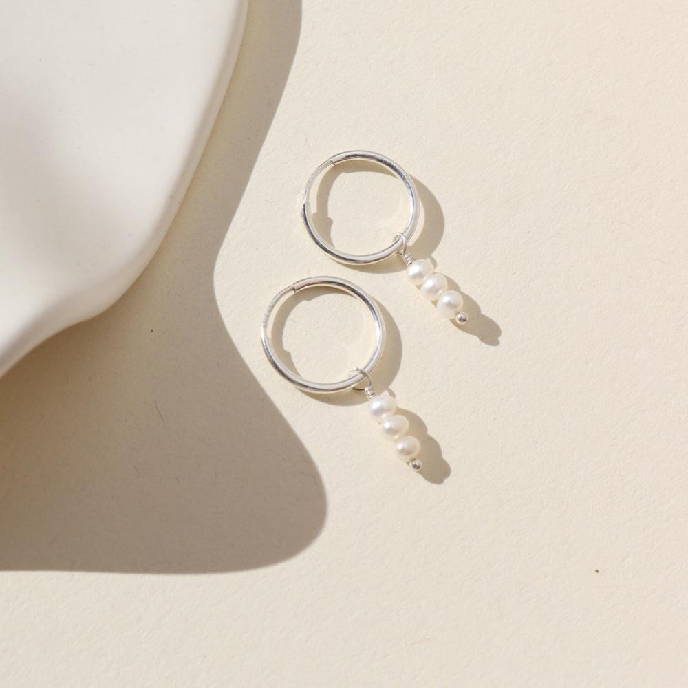 Silver petite hoops featuring three mini pearls hanging from each hoop. Earrings are displayed on cream backdrop. 