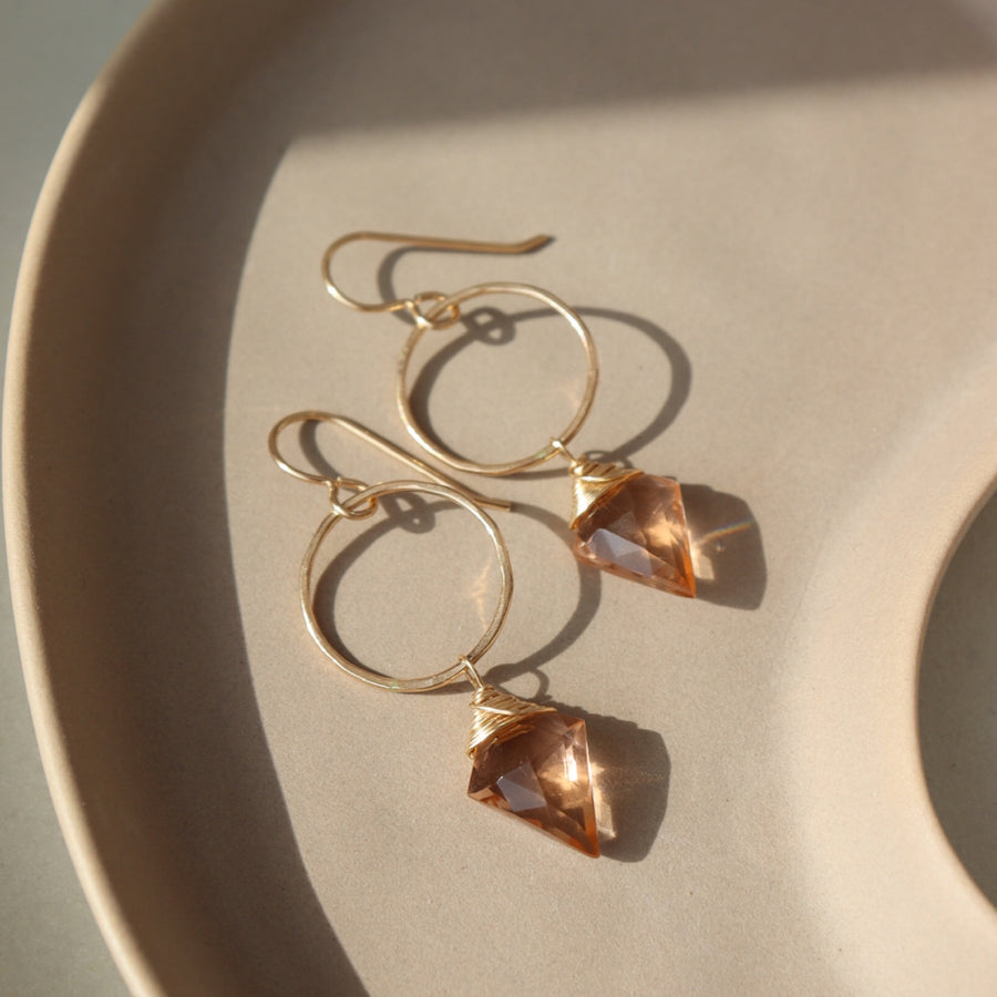 Champagne Quartz Drops laid on a peach colored plate laid in the sunlight. These earrings feature a hook earring with a open circle disc followed my the champagne quartz gemstone.