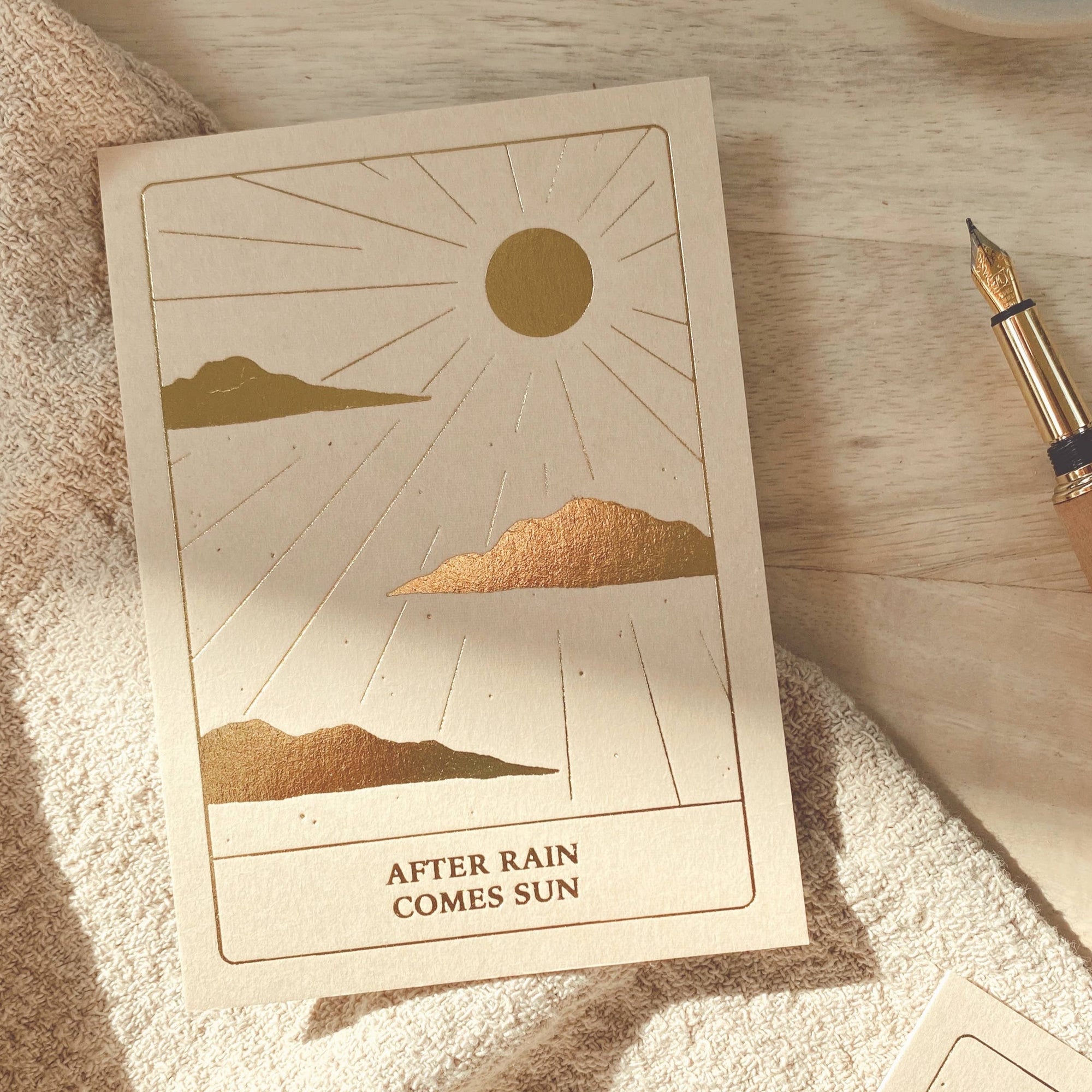 A card with a sun shining along with cloud on the bottom of the card it says "after the rain comes the sun." That is set on a blanket layer across the table.