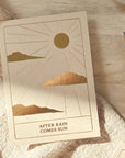 A card with a sun shining along with cloud on the bottom of the card it says "after the rain comes the sun." That is set on a blanket layer across the table.