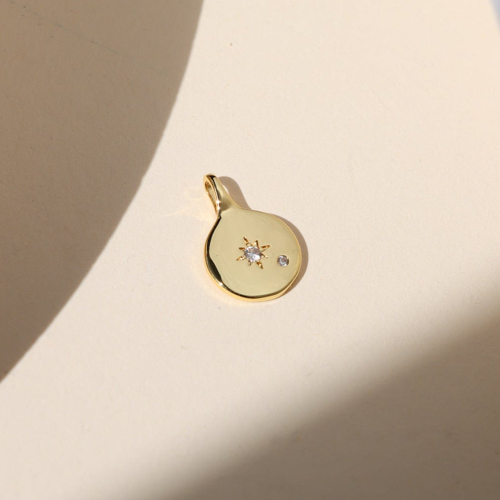 Gold coin charm with a starburst crystal detail and small accent stone, photographed on a warm neutral surface with soft natural light and shadows.