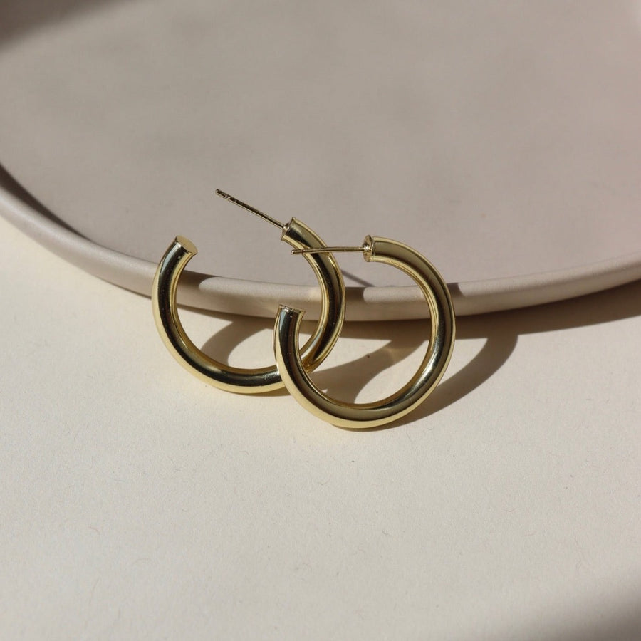 14k gold fill Harper Hoops laid on a tan paper in the sunlight. These earrings feature a classic smooth hoop with a post making the easy to take in and out.