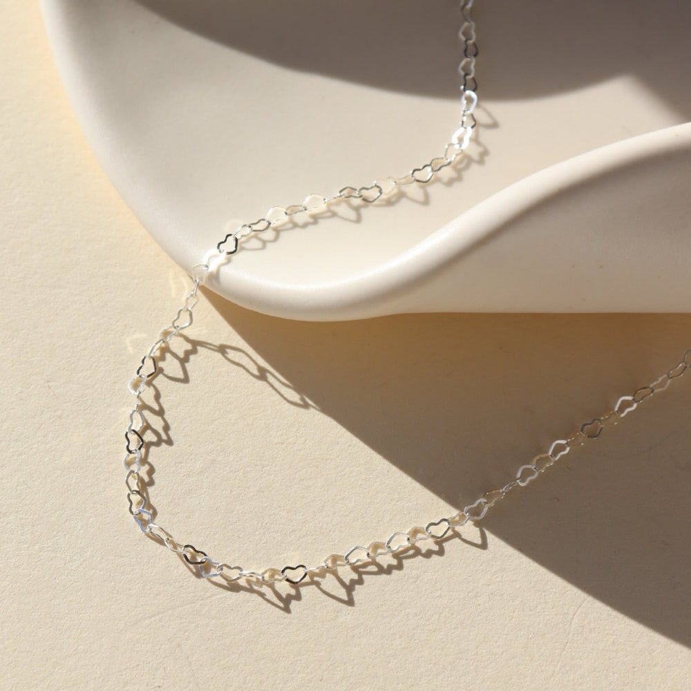 925 Sterling Silver Petite heart chain from Token Jewelry, photographed on a white, organically shaped ceramic dish. This chain features a silver tiny heart wire throughout the whole chain. 