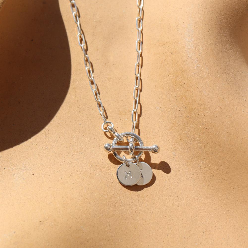 sterling silver monogram toggle necklace laid on a tan paper in the sunlight. This necklace features the narrow link chain connected by a toggle with the monogram disc. 