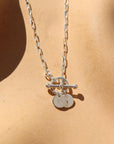 sterling silver monogram toggle necklace laid on a tan paper in the sunlight. This necklace features the narrow link chain connected by a toggle with the monogram disc. 