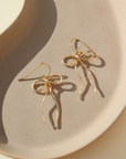 delicate 14k gold fill bow earring hand made by Token Jewelry, photographed on a ceramic dish in the sunlight