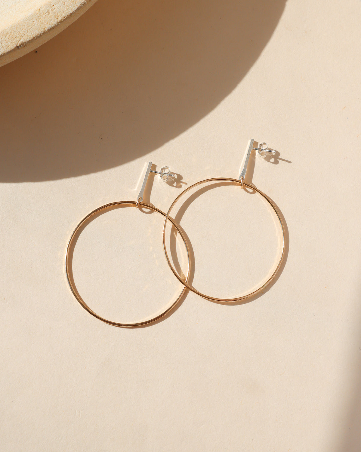 Gold hoop drop earrings with silver bar studs arranged on a cream background in warm natural sunlight.
